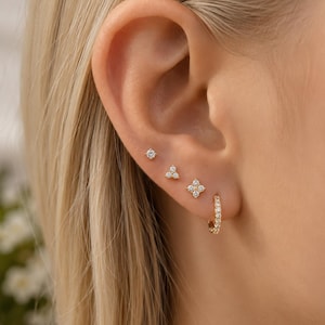 Könnte beinhalten: Ein Set aus vier goldenen Ohrringen, die an einem Ohr getragen werden. Die Kollektion umfasst einen kleinen Ohrstecker mit einem Stein, einen mit drei Steinen, einen blumenförmigen Stecker mit vier Steinen und einen kleinen goldenen Creolen-Ohrring.