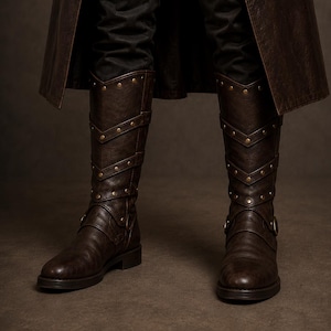 May include: A pair of dark brown knee-high boots featuring a textured finish, multiple horizontal straps with metallic studs arranged in a chevron pattern, and a buckled strap detail at the ankle, worn with black trousers and a long brown coat.