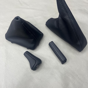 May include: Two black automotive gear shift boots with visible stitching, a black gear shift knob, and a black handbrake handle cover. These components are made of a smooth, matte material and are arranged together on a textured surface.