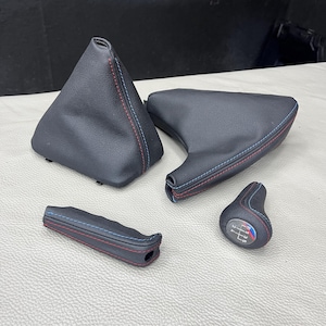Puede incluir: Un conjunto de componentes interiores de automóvil en color negro que incluye una funda para palanca de cambios, una funda para freno de mano, una empuñadura de freno de mano y un pomo de cambio manual con costuras rojas y azules.