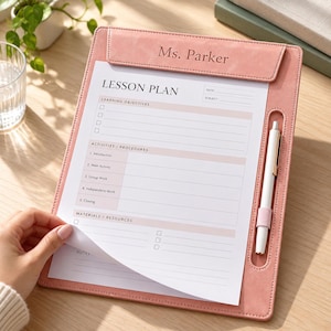 May include: A dusty rose faux leather clipboard with a top clip engraved with the name Ms. Parker. The clipboard holds a white lesson plan sheet with sections for learning objectives, activities, procedures, and materials. A white pen is held in a loop.