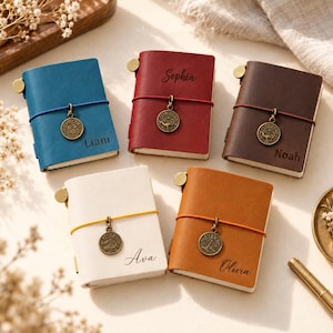May include: Five small leather journals in blue, red, dark brown, white, and tan. Each journal features a colored elastic band, a bronze-toned zodiac charm, and a personalized name engraved on the front cover: Liam, Sophia, Noah, Ava, and Olivia.