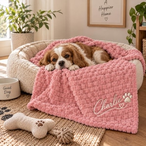May include: A plush, textured pink blanket with a waffle-knit pattern draped over a beige, round pet bed. The blanket features the embroidered name Charlie, a small heart, and a paw print. A brown and white dog rests inside the bed.