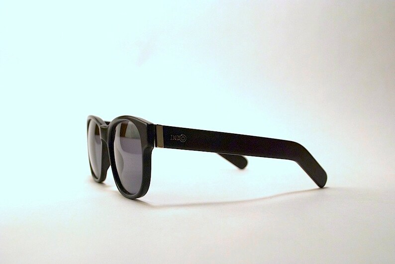 May include: Black sunglasses with a dark gray lens and a black frame. The frame has a silver logo that says "DXXO" on the right side.
