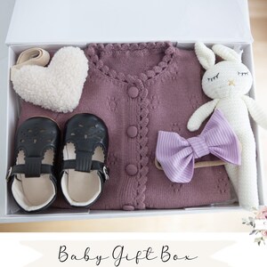 K&ouml;nnte beinhalten: Eine wei&szlig;e Geschenkbox gef&uuml;llt mit einem Baby-Geschenkset. Das Set enth&auml;lt einen lila gestrickten Cardigan, ein wei&szlig;es Pl&uuml;sch-Hasenspielzeug, ein Paar schwarze Schuhe, ein lila Schleifen-Haarband und ein wei&szlig;es, herzf&ouml;rmiges Pl&uuml;schspielzeug. Die Box ist mit "Baby Gift Box" und "OPTION 13" beschriftet.