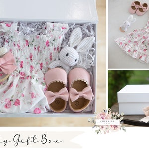 K&ouml;nnte beinhalten: Eine wei&szlig;e Geschenkbox gef&uuml;llt mit einem rosa Blumenkleid, rosa Schuhen, einem wei&szlig;en H&auml;kel-Hasen, einem rosa Stirnband und einem wei&szlig;en H&auml;kel-Hasen-Rassel. Die Box ist mit "Baby Gift Box" beschriftet.