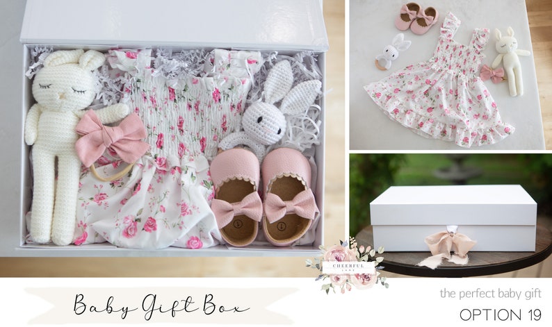 K&ouml;nnte beinhalten: Eine wei&szlig;e Geschenkbox gef&uuml;llt mit einem rosa Blumenkleid, rosa Schuhen, einem wei&szlig;en H&auml;kel-Hasen, einem rosa Stirnband und einem wei&szlig;en H&auml;kel-Hasen-Rassel. Die Box ist mit "Baby Gift Box" beschriftet.