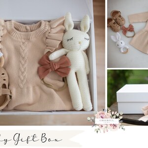 K&ouml;nnte beinhalten: Eine wei&szlig;e Geschenkbox gef&uuml;llt mit Babyartikeln, darunter ein Strickkleid, ein gestricktes Hasenspielzeug, ein Stirnband mit Schleife, ein Paar Schuhe und ein Holzring. Die Box ist mit "Baby Gift Box" beschriftet.