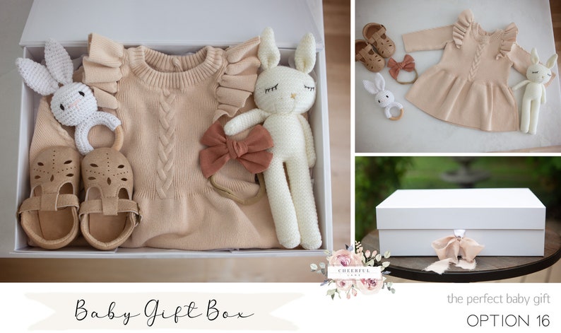 K&ouml;nnte beinhalten: Eine wei&szlig;e Geschenkbox gef&uuml;llt mit Babyartikeln, darunter ein Strickkleid, ein gestricktes Hasenspielzeug, ein Stirnband mit Schleife, ein Paar Schuhe und ein Holzring. Die Box ist mit "Baby Gift Box" beschriftet.