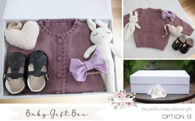K&ouml;nnte beinhalten: Eine wei&szlig;e Geschenkbox gef&uuml;llt mit einem Baby-Geschenkset. Das Set enth&auml;lt einen lila gestrickten Cardigan, ein wei&szlig;es Pl&uuml;sch-Hasenspielzeug, ein Paar schwarze Schuhe, ein lila Schleifen-Haarband und ein wei&szlig;es, herzf&ouml;rmiges Pl&uuml;schspielzeug. Die Box ist mit "Baby Gift Box" und "OPTION 13" beschriftet.