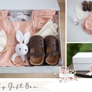 K&ouml;nnte beinhalten: Eine wei&szlig;e Geschenkbox gef&uuml;llt mit einem rosafarbenen Baby-Outfit, einem Paar brauner Schuhe, einem wei&szlig;en H&auml;kel-Hasen, einem Holzring und einem wei&szlig;en Stirnband mit Schleife. Die Box ist mit "Baby Gift Box" und "OPTION 25" beschriftet.