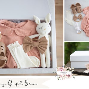 K&ouml;nnte beinhalten: Eine wei&szlig;e Geschenkbox gef&uuml;llt mit Babyartikeln, darunter ein rosa Strampler, wei&szlig;e Socken, braune Schuhe, ein Holzring und zwei geh&auml;kelte Hasenspielzeuge. Die Box ist mit einem braunen Band gebunden und der Text "Baby Gift Box" steht auf der Vorderseite.