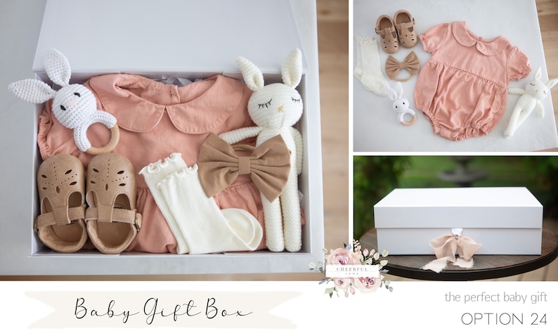 K&ouml;nnte beinhalten: Eine wei&szlig;e Geschenkbox gef&uuml;llt mit Babyartikeln, darunter ein rosa Strampler, wei&szlig;e Socken, braune Schuhe, ein Holzring und zwei geh&auml;kelte Hasenspielzeuge. Die Box ist mit einem braunen Band gebunden und der Text "Baby Gift Box" steht auf der Vorderseite.