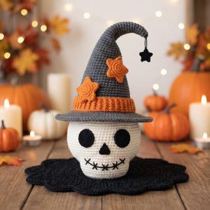 Peut inclure: Un crâne crocheté portant un chapeau pointu gris avec un bord orange et des appliqués en forme d'étoiles orange. Le chapeau présente une petite étoile noire suspendue à la pointe. Le crâne a des yeux ronds noirs, un nez triangulaire et une bouche cousue.