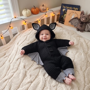 May include: A black crocheted bat costume for an infant, featuring a hood with pointed ears, long sleeves, and attached gray bat wings. The garment includes three black toggle buttons down the front and gray cuffs at the ankles, creating a complete bat-themed outfit.