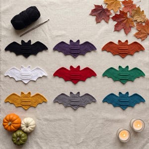 May include: Nine crocheted bat appliques in black, purple, orange, white, red, green, yellow, gray, and blue. A ball of black yarn and a crochet hook are positioned at the top left, with three small pumpkins and two tea light candles below.