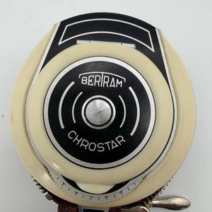 May include: A round, cream-colored Beriram Chrostar light meter with a black circular center featuring white text and curved lines. The device has a silver metallic button in the middle and a graduated scale along the bottom edge, partially encased in a dark red textured leather case with a silver zipper.