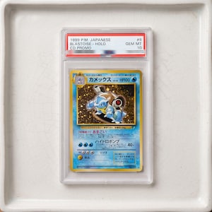 May include: A 1999 Japanese Blastoise holographic trading card encased in a clear protective slab. The card features the character Blastoise with a gold border, blue background, and Japanese text. The label at the top reads 1999 P.M. JAPANESE BLASTOISE - HOLO CD PROMO #9 GEM MT 10.