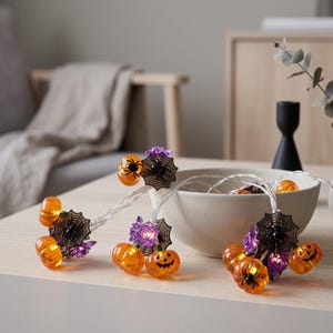 May include: A string of Halloween decorative lights featuring orange jack-o'-lantern pumpkins, black spider web ornaments with small black spiders, and purple flower accents. The lights are connected by a clear wire and are partially coiled in a bowl.