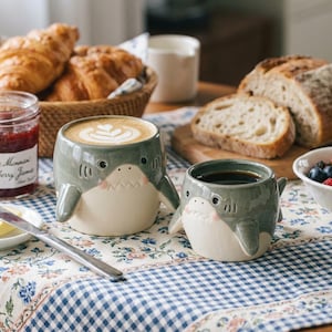 Peut inclure: Deux tasses en céramique en forme de requins gris avec des ventres blancs, de petites nageoires et des traits de visage peints. La plus grande tasse contient un latte avec de la mousse, tandis que la plus petite contient du café noir.