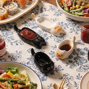May include: A collection of ceramic cat-shaped sauce dishes in black and white, featuring elongated bodies with tails and small ears. These small bowls are designed to hold condiments and are arranged on a floral patterned tablecloth.