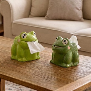 Op de afbeelding: Twee keramische tissuehouders in de vorm van een kikker in verschillende tinten groen. Elke kikker heeft grote witte ogen met zwarte pupillen, roze wangvlekken en een brede mondopening waaruit een wit tissue in het midden naar buiten komt.