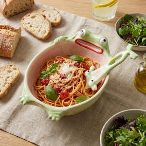 Puede incluir: Un cuenco de cerámica de color verde claro con forma de rana, con la boca muy abierta y una cuchara de servir a juego con forma de rana. El cuenco contiene espaguetis con salsa de tomate roja, queso rallado y hojas de albahaca fresca.
