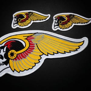 Könnte beinhalten: Drei gestickte Aufnäher mit einem weißen Totenkopf, der einen geflügelten Helm mit gelben und roten Federn trägt. Das Design umfasst einen großen mittleren Aufnäher und zwei kleinere passende Aufnäher, alle mit weißem Garn auf schwarzem Grund.