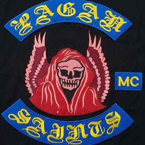 May include: A black fabric patch set featuring a central red hooded skull figure with pink wings. Above is a blue arched patch with yellow text reading PAGAN. Below is a blue arched patch with yellow text reading SAINTS. A small blue rectangle with yellow MC text is on the right.