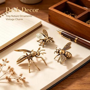 May include: Three brass-toned metal insect ornaments shaped like bees and a wasp, featuring detailed wings and legs. The text "Desk Decor" and "Tiny Nature Ornaments Vintage Charm" is visible on the page. These metallic figures are arranged on a notebook.