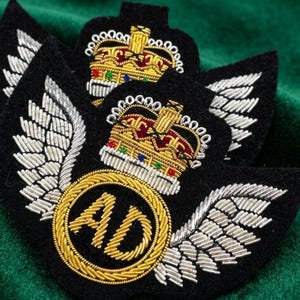 Può includere: Una toppa in tessuto nero con un intricato ricamo in filo d'oro e d'argento. Il design presenta un cerchio centrale dorato con le lettere AD, fiancheggiato da ali argentate e sormontato da un dettagliato emblema di corona reale in oro, rosso e blu.
