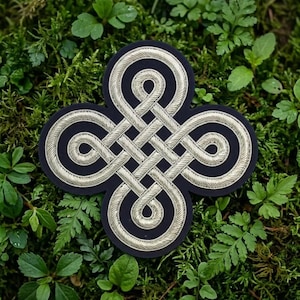 May include: A dark navy blue patch featuring a metallic silver embroidered Celtic knot design. The embroidery consists of intricate, continuous interlacing lines forming a symmetrical four-lobed shape with a central woven pattern.