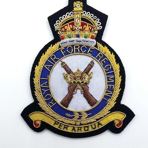 May include: An embroidered patch featuring the Royal Air Force Regiment emblem. It displays a central white circle with crossed rifles and a crown, surrounded by a blue ring with gold text, topped with a large crown, and a gold banner reading PER ARDUA.