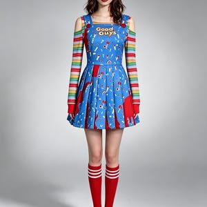 May include: A blue overall-style dress featuring the text Good Guys, a colorful pattern of small shapes, and red accents. It is worn over a long-sleeved shirt with horizontal rainbow stripes and cold-shoulder cutouts, paired with red knee-high socks.