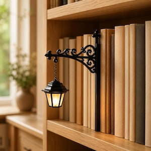 May include: A black metal bookend featuring an ornate, scrollwork bracket that extends outwards to support a miniature, hanging lantern. The lantern is suspended by a small chain and emits a soft, warm glow from within its glass panels.