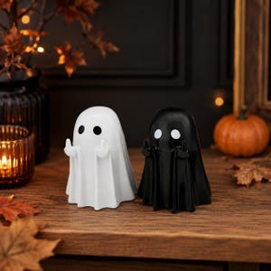 Peut inclure: Deux figurines de fantômes, une blanche et une noire, chacune dotée de deux yeux ronds et de bras levés avec les majeurs tendus. Les figurines lisses et brillantes ont la forme de draps drapés, posées côte à côte comme objets décoratifs.