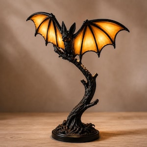 May include: A table lamp shaped like a black bat perched on a textured tree branch. The bat has outstretched wings with translucent, glowing amber-coloured panels that emit a warm light, supported by a dark, sturdy base resembling tree roots.