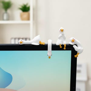 Könnte beinhalten: Vier kleine weiße Gänsefiguren mit orangefarbenen Schnäbeln und Füßen sitzen auf der Oberkante eines schwarzen Computermonitors. Jede Figur nimmt eine andere Pose ein, darunter liegend, kopfüber hängend, aufrecht sitzend und über die Ecke gelehnt.