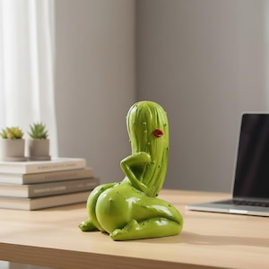 May include: A green, anthropomorphic pickle figurine in a kneeling pose. The figure features a textured, bumpy surface, a single arm resting on its hip, and prominent red lips. The sculpture is rendered in a glossy finish with a distinct, curved shape.