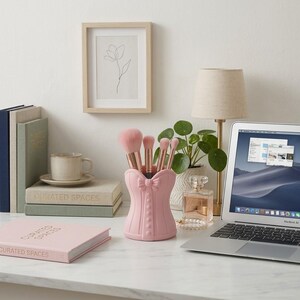Peut inclure: Un porte-pinceaux de maquillage rose clair en forme de corset avec un nœud central et des boutons, contenant quatre pinceaux à maquillage aux manches roses. Le support est posé sur une surface blanche près de livres, d'une tasse et d'un ordinateur.