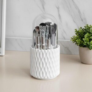 Peut inclure: Un organisateur de pinceaux de maquillage cylindrique blanc avec un motif de vagues verticales texturé et un couvercle en plastique transparent en forme de dôme. Le récipient contient divers pinceaux et tubes, maintenus droits et visibles.