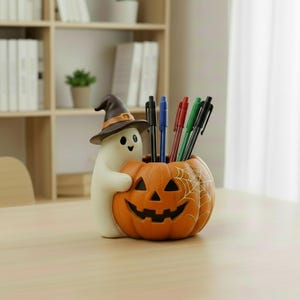 Könnte beinhalten: Ein Schreibtisch-Organizer in Form eines weißen Geistes mit braunem Hexenhut, der einen orangefarbenen Kürbis mit weißem Spinnennetz-Muster umarmt. Der Kürbis enthält fünf Stifte in den Farben Schwarz, Blau, Rot und Grün auf einer Fläche.