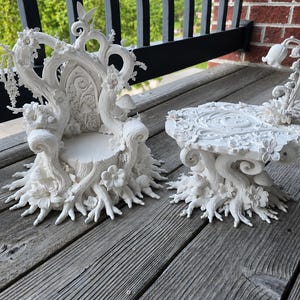 May include: A white miniature chair and table set featuring intricate sculpted details of tree roots, branches, flowers, and mushrooms. The chair has a high back with a butterfly at the top, while the table features a flat surface with floral accents and a curved stem support.