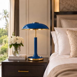 May include: A table lamp with a glossy blue dome-shaped shade and a gold-toned metal base. The lamp features a central cylindrical stem and a circular base. It is positioned on a dark wooden nightstand next to a glass vase of white flowers.