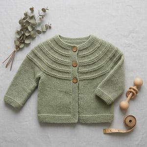 Könnte beinhalten: Eine salbeigrüne Strickjacke mit Rundhalsausschnitt, langen Ärmeln und drei braunen Holzknöpfen auf der Vorderseite. Das Kleidungsstück hat ein strukturiertes Kraus-rechts-Muster an der Passe und den Bündchen sowie einen glatten Rumpf.