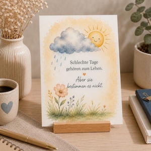 Può includere: Un biglietto d'auguri rettangolare con un'illustrazione ad acquerello di un sole sorridente, una nuvola di pioggia e un campo di fiori. Il testo in tedesco recita: 'Schlechte Tage gehören zum Leben. Aber sie bestimmen es nicht.' su un supporto in legno.