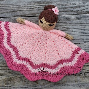 May include: A handmade crochet baby lovey in shades of pink, featuring a doll head with brown hair and a flower. The lovey has a scalloped edge and is designed to comfort and soothe infants. The arms are tan.