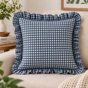 Könnte beinhalten: Ein quadratisches Zierkissen mit einem blau-weißen Vichy-Karo-Muster. Das Kissen ist mit einem Rüschenrand aus demselben blau-weiß karierten Stoff versehen, der eine dekorative Kante um den gesamten Umfang des Kissens bildet.