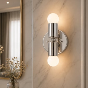 May include: A wall-mounted light fixture featuring a polished chrome finish with a circular backplate and two cylindrical sockets. Two spherical, frosted white glass bulbs are positioned at the top and bottom, emitting a warm, soft glow.