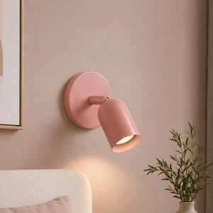 Puede incluir: Un aplique de pared de color rosa mate con una base circular y un cabezal cilíndrico. La lámpara está colocada para emitir una luz cálida y enfocada hacia abajo, con un diseño sencillo y limpio que se fija directamente a la pared.